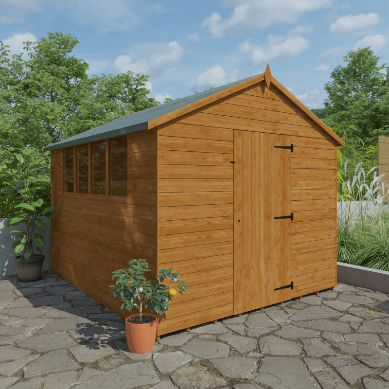 Wooden shed with a green roof on a stone patio with plants and trees in the background