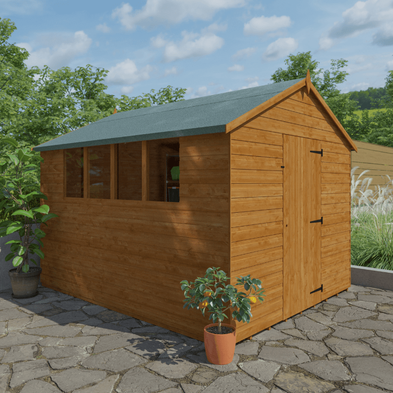 Wooden shed with a green roof on a stone patio with plants around, surrounded by trees and blue sky.