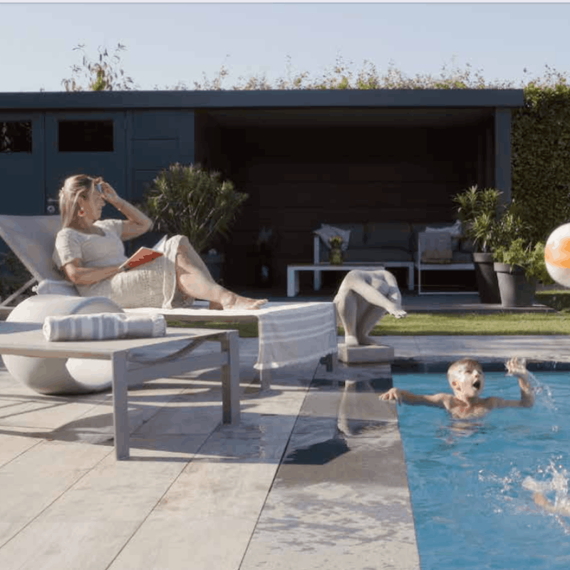 Woman reading a book on a lounge chair by a pool with a child playing in the water.